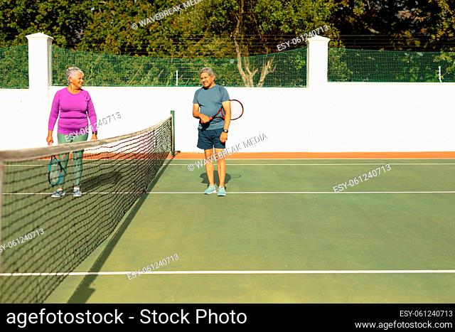 Biracial senior man talking with senior wife while standing by net at tennis court on sunny day