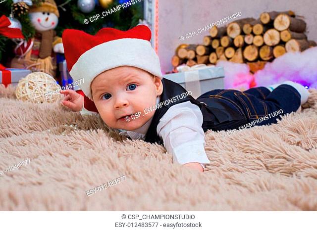 Baby in Santa Claus hat lying on the carpet. Christmas tree in the background