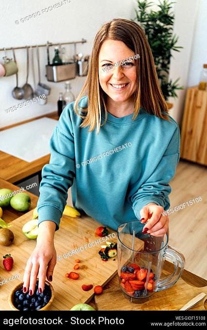 Happy blond woman with fresh fruits in blender standing at table in kitchen