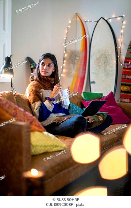 Young woman in knit sweater relaxing on living room sofa with drink