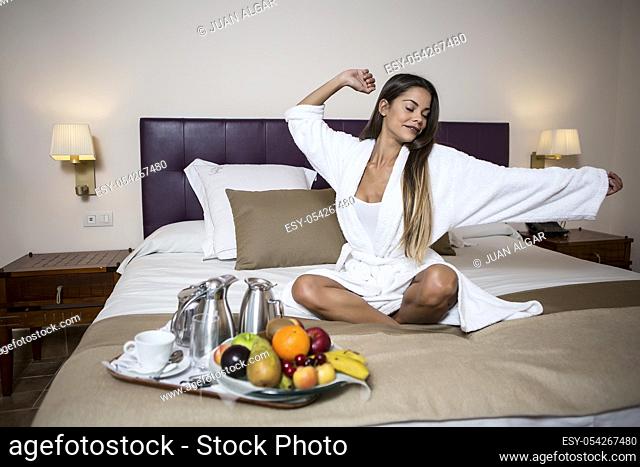 Young pretty woman sitting on bed in morning gown near plate of fresh fruits and coffee set and stretching with closed eyes