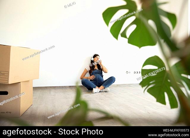 Pregnant woman listening music through headphones while sitting against wall in new home