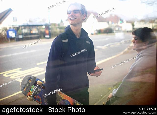 Happy teenage boy with smart phone and skateboard at bus stop
