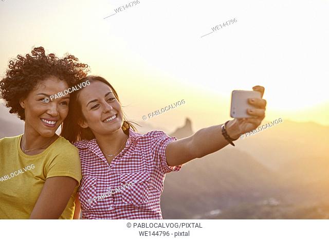 Two girl friends taking a selfie with mountains behind them while sitting on a stone wall and wearing casual clothing