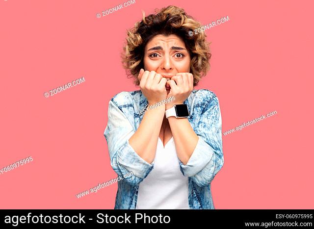 Portrait of nervous young woman with curly hairstyle in casual blue shirt standing, bitting her nails and looking at camera with panic face expression