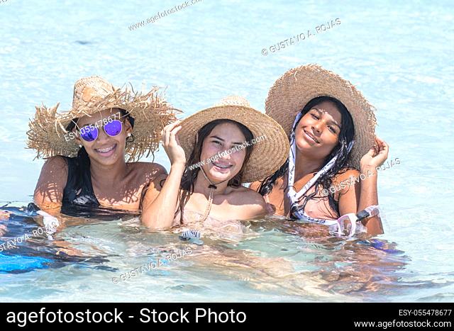 Girls multiracial friends with hat enjoying sitting inside water in sunny day at Los Roques Venezuela