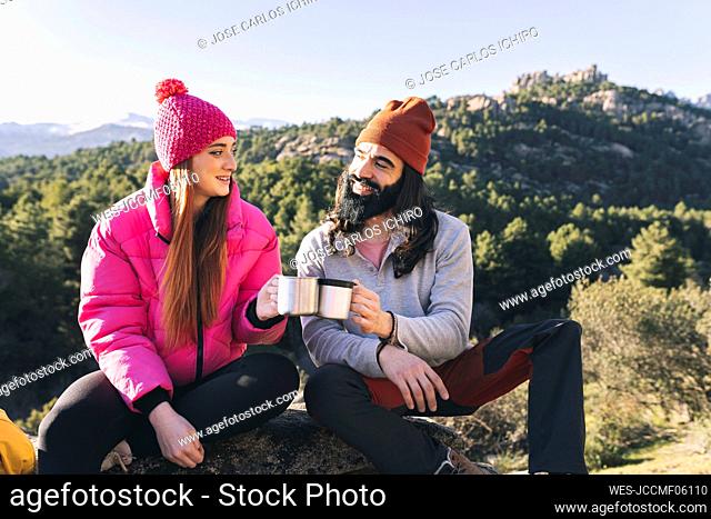Smiling man and woman toasting mugs sitting on rock