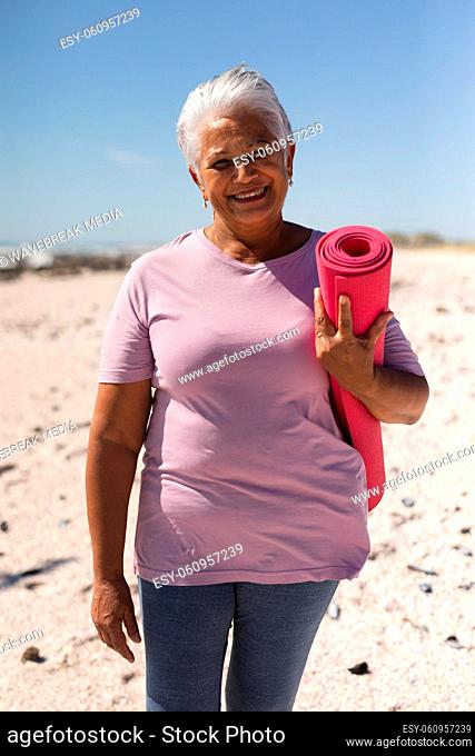 Portrait of smiling retired senior biracial woman holding yoga mat with short white hair at beach
