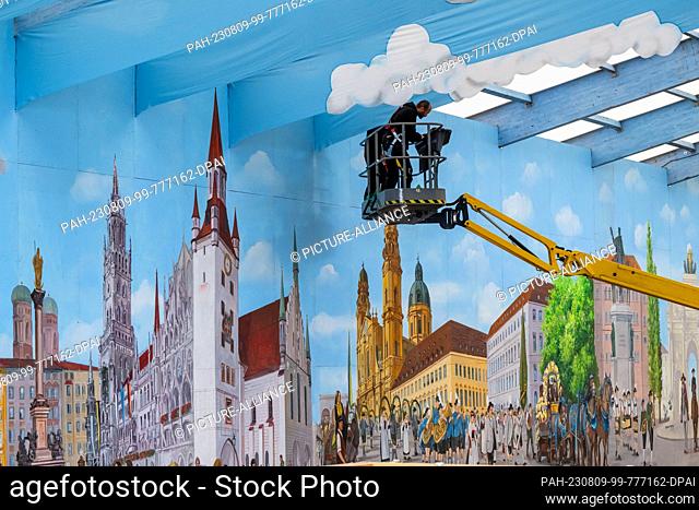 09 August 2023, Bavaria, Munich: Two workers on a lifting platform attach blue fabric panels with a cloud pattern under the roof structure of a beer tent on the...