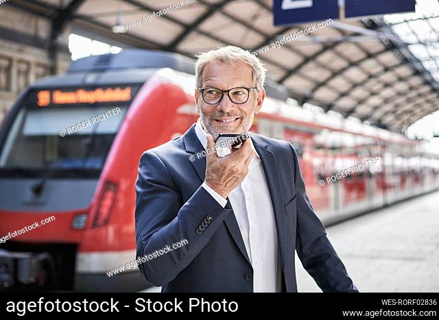 Smiling male professional looking away while sending voicemail through smart phone on railroad station