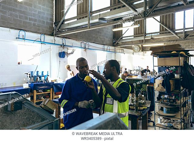 Front view of two young African American male colleagues in discussion beside a machine at a cricket ball factory, one is holding a yellow ball