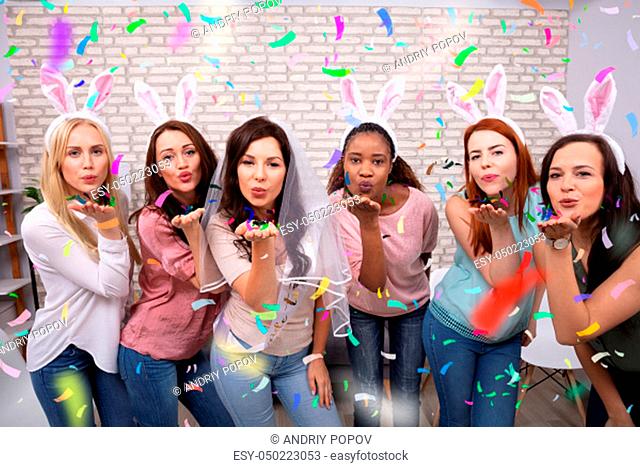 Close-up Of Attractive Female Friends Blowing Paper Confetti Towards Camera Standing Against Brick Wall