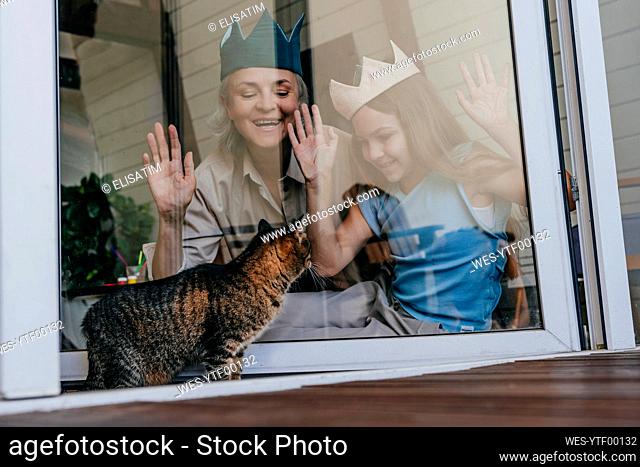 Happy grandmother and granddaughter wearing crown looking at cat through glass window