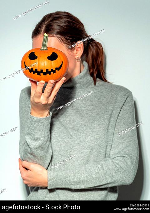 Crop unrecognizable woman holding halloween scary face bright orange pumpkin in front of her face. Young woman in gray cashemere sweater hides her face behind...