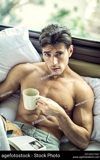Sexy handsome young man laying shirtless on his bed next to window, holding a coffee or tea cup