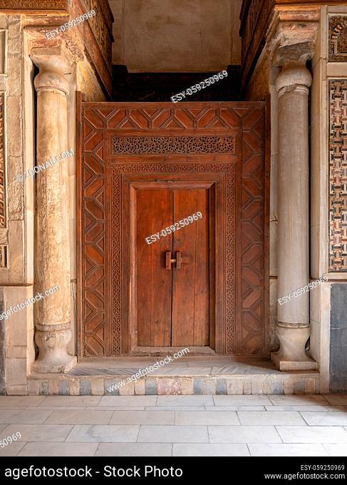 Old wooden door framed by wooden engraved panels decorated with geometric and floral patterns between two carved columns located at Sultan Qalawun Mausoleum