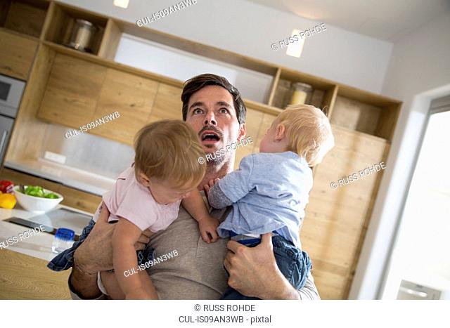 Father struggling to carry male and female twin toddlers in dining room
