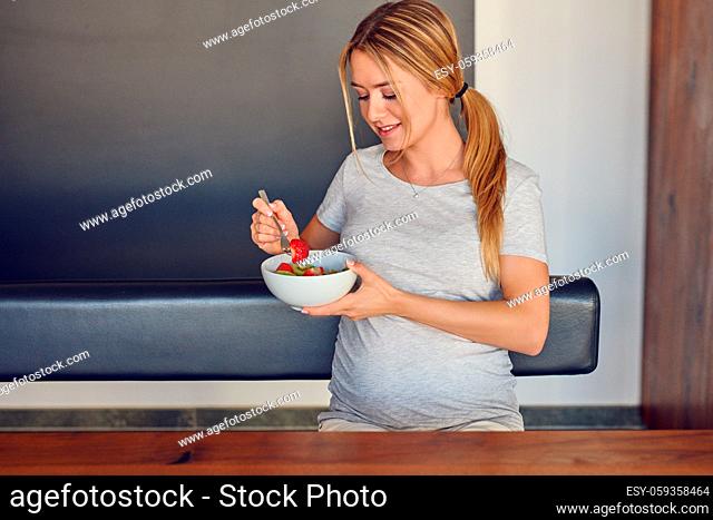 Young pregnant woman enjoying a healthy fresh fruit salad smiling happily at the camera as she holds the bowl in her hand taking a mouthful seated at a table
