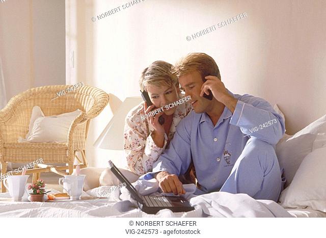 portrait, indoor, full-figure, young blond couple wearing pyjama sit with a notebook at breakfast in bed, both making a call with a mobile phone  - GERMANY