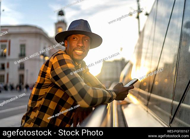 Smiling african man with smart phone looking away while standing in city during sunset