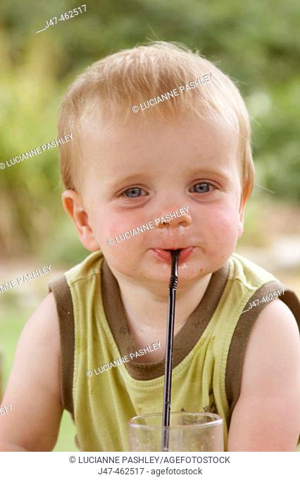 1 year old baby boy smiling cheekily into camera, outside on a sunny day, with a straw in his mouth