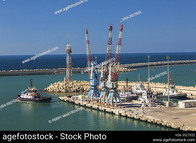 Tugboat approching dock with four-link cargo loading cranes and docked tugboats, Ashdod Port, Israel