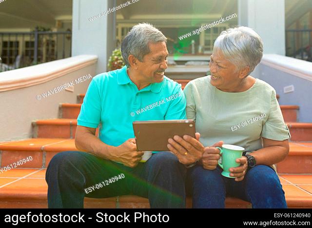 Smiling biracial senior man holding digital pc talking with wife holding cup while sitting on steps