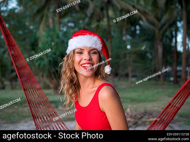 Young happy woman in santa hat and red swimsuit showing her tongue while sitting on the hammock on the sandy beach. Christmas beach vacation travel woman...