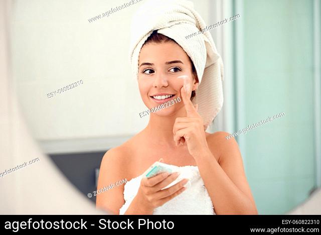 Pretty young woman in white towels in a bathroom giving the camera a lovely friendly smile as she applies cream to her cheeks while bathing in a skin care and...