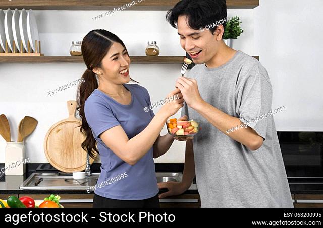 Asian couple spend time together in the kitchen. Young woman feed her boyfriend a piece of apple from salad bowl