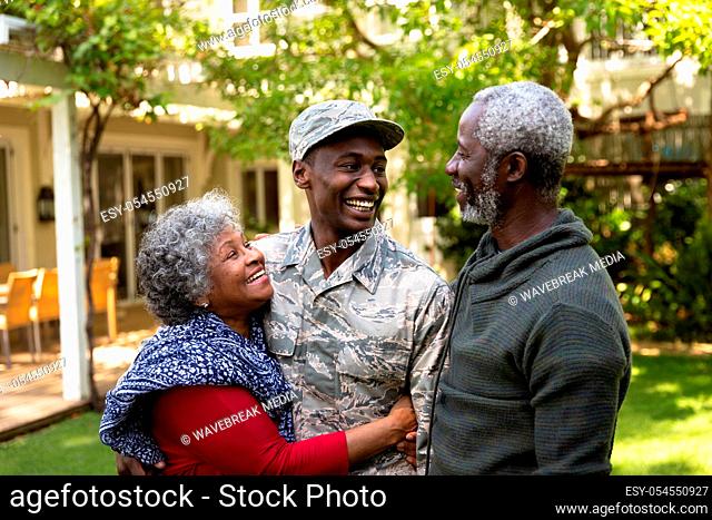 Front view of a young adult African American male soldier in the garden outside his home embracing and smiling with his parents