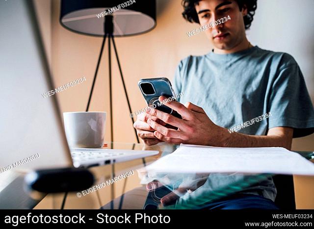 Man using mobile phone while sitting with laptop at table