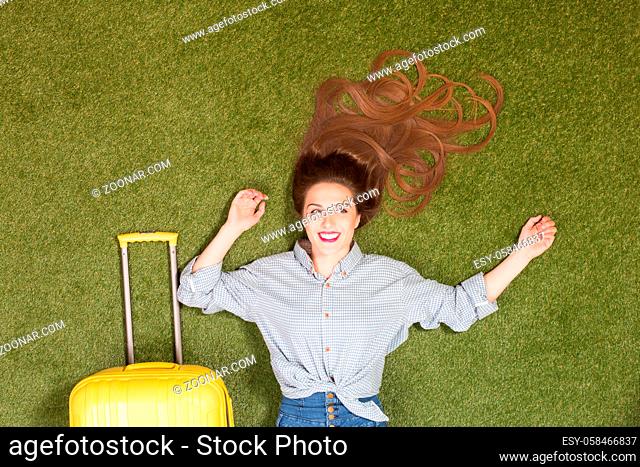 Portrait of beautiful tourist lady lying on green grass. Pretty lady with long brown hair lying near yellow luggage. Ready for travelling, trip or journey