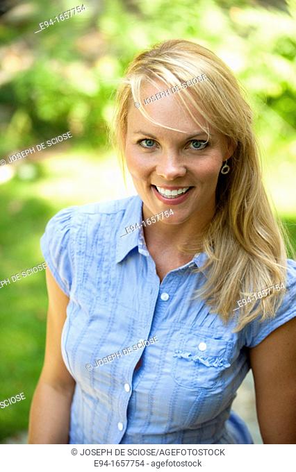 Portrait of a 35 year old blond woman wearing a blue blouse and jeans in a garden setting looking at the camera