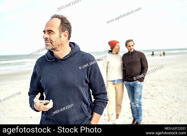 Portrait of man standing on sandy beach with smart phone in hand with young couple in background