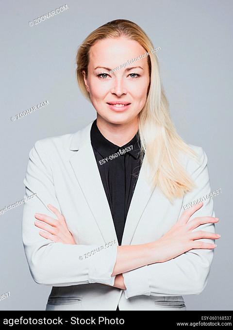 Portrait of cheerful, beautiful, smart, young businesswoman in white business attire, standing with arms crossed against gray background