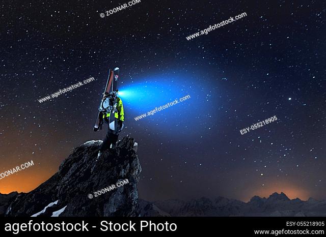 Night landscape. A professional backcountry skier with a backpack and skis stands on a rock in the mountains and shines headlamp into the sky