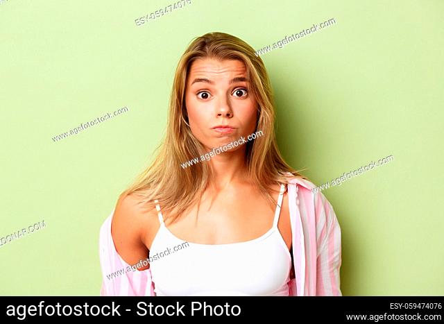 Close-up of silly blond girl in pink shirt, grimacing and looking confused, standing over green background