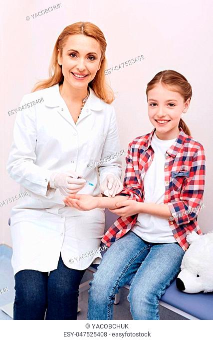 Give your hand. Positive delighted girl sitting near nurse and white bear holding her right hand while looking forward