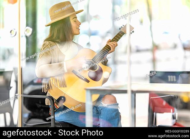 Young female musician playing guitar seen through glass at cafe