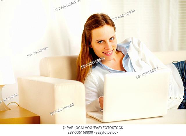 Portrait of a attractive caucasian woman lying on sofa with a laptop and looking at you