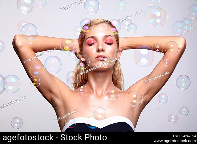 Beautiful young blonde woman with soap bubbles on grey background. Female with eyes closed with hands up