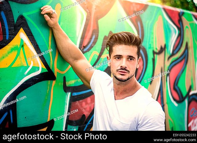 Attractive muscle man leaning on colorful graffiti wall, wearing white t-shirt