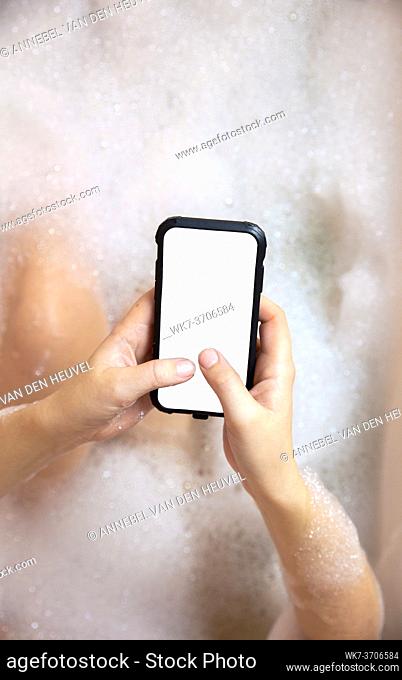 young female holding blank mobile phone, smart phone in bathtub with soapy foam close-up top view background copy space