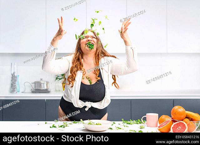 Happy chubby girl scattering spinach leaves all over the table. Overweight girl happy loosing weight eating fresh salad. Curvy body young woman with long blond...