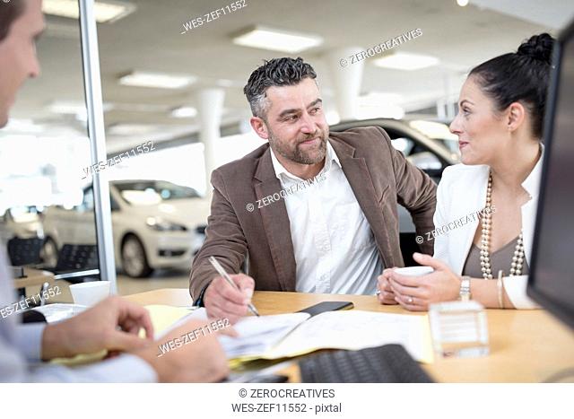 Car dealer and couple sitting at desk with agreement forms in showroom