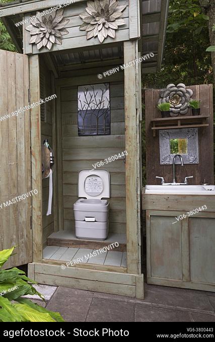 Wood plank outhouse with manual flush portable toilet and wooden cabinet with sink in backyard, Quebec, Canada