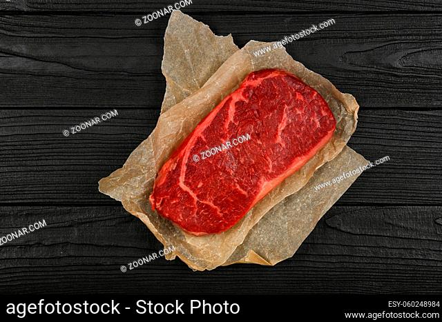 Close up one aged prime marbled raw sirloin beef steak on brown paper parchment wrapping over black wooden table background, elevated top view, directly above