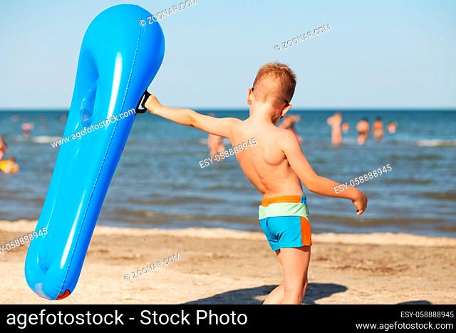 Little kid holding an inflatable mattress on the beach on hot summer day. Smiling boy playing on the beach with air mattress