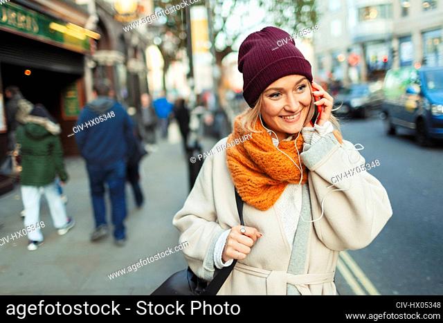 Young woman in stocking cap and scarf talking on smart phone on urban sidewalk